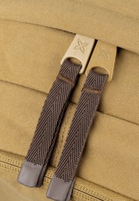 Detailný pohľad na khaki látkový batoh s dvoma hnedými zipsami, ktoré majú pozlátené kovové prvky a textúrované úchytky.