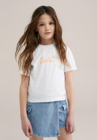 Camiseta de algodón blanca con el logo de "Barbie" en melocotón, combinada con una falda de mezclilla clara con bolsillos laterales y un dobladillo curvado.