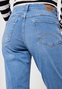 Jeans de mezclilla azul claro con cintura alta, que presentan dos bolsillos traseros y una etiqueta de piel marrón. La tela tiene una textura suave y una costura clásica.