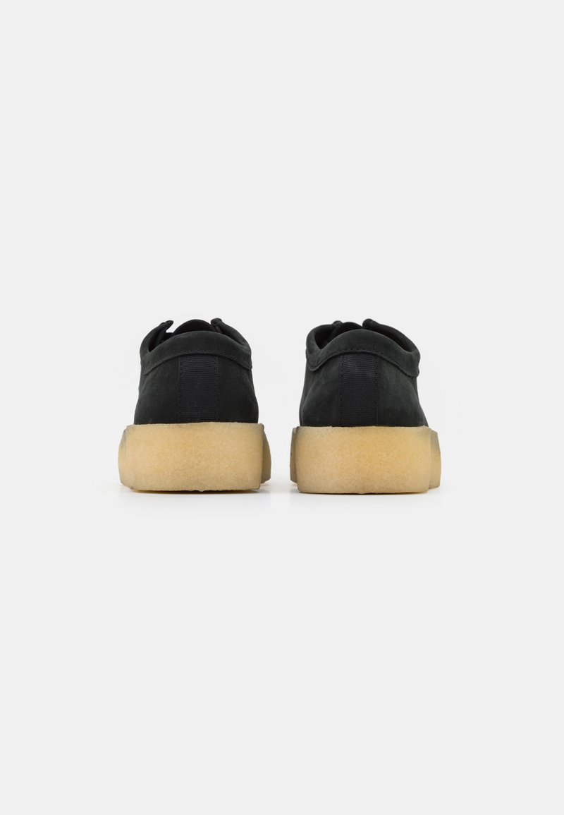 Sneakers neri in suede con profilo basso, caratterizzati da una parte superiore morbida e testurizzata e una suola in gomma color tan a contrasto. La vista posteriore mostra i dettagli delle cuciture.