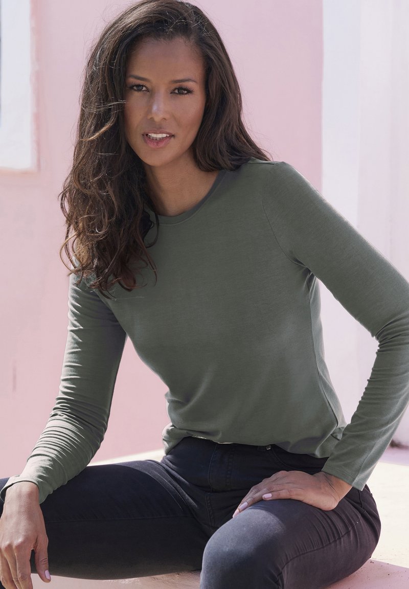 Olijf groene top met lange mouwen, gemaakt van zachte, lichte stof; heeft een ronde halslijn en een getailleerde pasvorm. Gemaakt in combinatie met donkere jeans.