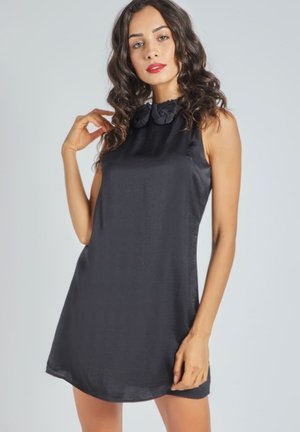 Mujer con cabello rizado largo lleva un vestido mini negro sin mangas con detalle floral en el collar, posando frente a un fondo gris claro.