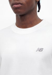 Prim-plan cu o persoană care poartă un tricou alb cu guler rotund și un mic logo gri New Balance pe pieptul stâng.