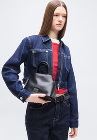 Γυναίκα ντυμένη με σκούρο τζιν μπουφάν και παντελόνι, κόκκινο τοπ, κρατώντας μικρή μαύρη τσάντα με μεταλλικό κούμπωμα μπροστά σε απλό φόντο.