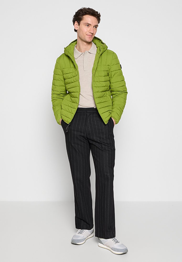 Giacca puffer trapuntata verde con cappuccio, polo beige, pantaloni neri a righe sottili e sneakers grigio chiaro.