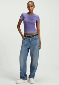 Camiseta ajustada de color púrpura claro con texto, combinada con unos jeans azules de gran tamaño, con un cinturón negro y zapatos blancos gruesos.