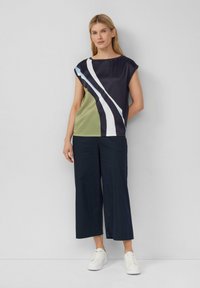 Bluse mit kurzen Ärmeln in Marineblau, Olive und Weiß mit kräftigen diagonalen Streifen. Kombiniert mit weit geschnittenen marineblauen Hosen und weißen Sneakern.