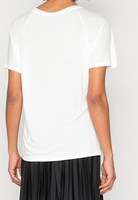 Camiseta blanca de manga corta con cuello redondo, textura suave y un corte relajado, vista desde la espalda, combinada con una falda negra con plisados.