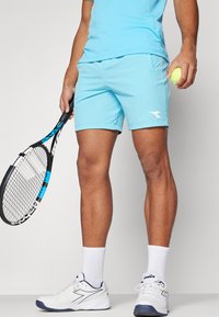 Heledad helesinised sportlikud lühikesed püksid koos valge logoga, valged sokid ja tennisetossud. Tennisereket ja pall on käes.