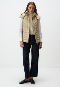 Beige gesteppte Weste mit Kapuze, mit einem Reißverschluss vorne und Rautenmuster. Getragen über einem cremefarbenen Pullover und kombiniert mit dunklen Jeans und schwarzen Slippern.