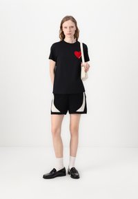 T-shirt noir avec un graphique de cœur rouge, associé à un short noir avec des motifs abstraits crème. Le mannequin porte un petit sac crème et des chaussures noires.