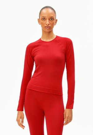 Vrouw draagt een aansluitende rode lange mouw top en bijpassende rode broek, staand tegen een effen witte achtergrond.