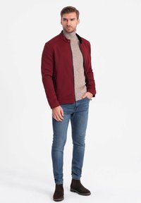 Burgundy zip-up jacka i strukturerad tyg, tillsammans med en ljusbrun polo, blå tajta jeans och mörkbruna ankelstövlar.