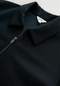 Black collared shirt with partially zipped silver zipper and visible label inside the neckline reading "N. Premium Collection."