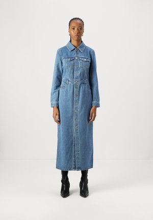 Lys blå denimkjole med lange ermer, knappestolpe foran, to brystlommer og belte i livet; faller til midt på leggen. Brukt med svarte støvler.