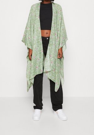 Persona con un kimono floral verde claro sobre un top corto negro, pantalones negros y zapatillas blancas sobre un fondo liso.