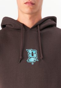 Detailný záber na osobu, ktorá má na sebe tmavo hnedú mikinu s kapucňou, na hrudi s vyšitým modrým stilizovaným logom "hur" a so šnúrkami visiacimi nadol.