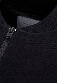 Svart zip-up jacka med en slät textur, utrustad med dragkedja, ribbad krage och en etikett med texten "JACK & JONES CORE."