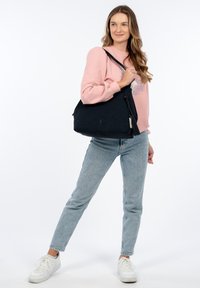 Borsa a tracolla in tessuto nero con motivo testurizzato, chiusura a zip e dettaglio con frange. Il modello indossa jeans azzurri e un maglione rosa.