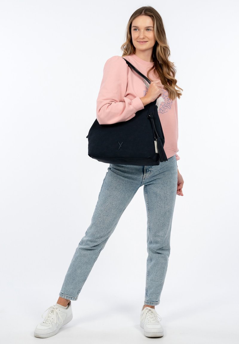 Borsa a tracolla in tessuto nero con motivo testurizzato, chiusura a zip e dettaglio con frange. Il modello indossa jeans azzurri e un maglione rosa.
