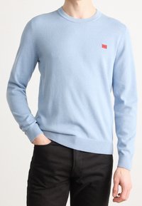 Pullover azzurro chiaro a maglia con collo rotondo, polsini e orlo a coste, caratterizzato da un piccolo etichetta rossa sul petto. Abbinato a pantaloni neri.