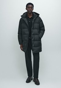Uomo in piedi con indosso un lungo giubbotto a sacco nero, una polo nera, pantaloni neri e scarpe di pelle nere su uno sfondo chiaro e uniforme.
