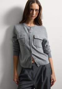 Cardigan gris à boutons en tissu tricoté doux. Comprend deux poches avant et des poignets côtelés. Associé à un pantalon gris taille haute.