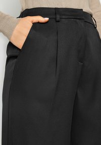 Zwarte op maat gemaakte broek met voorzakken, met een gladde textuur en een gestructureerd ontwerp. De tailleband heeft riemlussen voor extra detail.