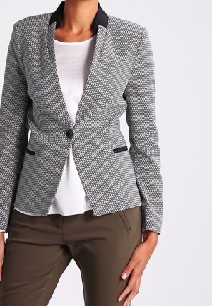 Mujer con un blazer ajustado de patrón blanco y negro sobre una camiseta blanca y pantalones marrones, de pie con una mano en la cadera.
