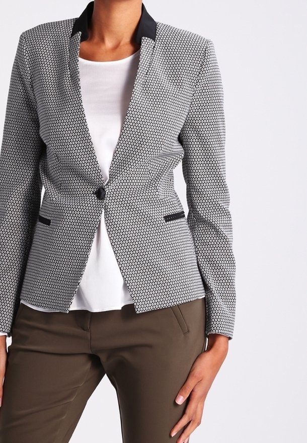 Femme portant un blazer ajusté blanc et noir à motifs sur un haut blanc et un pantalon marron, debout avec une main sur la hanche.