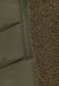Tessuto intrecciato marrone e materiale imbottito verde oliva uniti da una cerniera; presenta una texture morbida e un contrasto di colori distintivo.