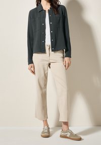 Dunkelgrünes, strukturiertes Hemd mit Knöpfen und zwei Brusttaschen, getragen über einem weißen T-Shirt, kombiniert mit hellbeigen Hosen und hellen Sneakers.