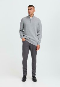 Grijze zip-up trui van zacht materiaal, gecombineerd met donkergrijze slim-fit broek en zwarte schoenen. Model staat met de handen in de zakken, neutrale achtergrond.