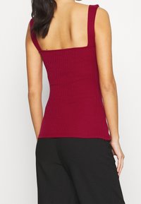 Donna che indossa un top ribattuto senza maniche color bordeaux e pantaloni neri larghi, mostrata di schiena su uno sfondo bianco uniforme.