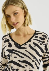 Suéter beige con cuello en V y estampado abstracto de animal en negro, con un borde texturizado negro con brillo a lo largo del escote. Material de punto suave.