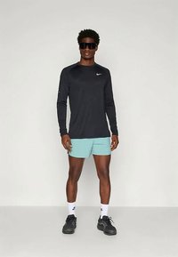 Μαύρο αθλητικό μπλουζάκι με μακριά μανίκια και λευκό λογότυπο Nike, συνδυασμένο με ανοιχτό μπλε σορτς. Μαύρα αθλητικά παπούτσια και γυαλιά ηλίου ολοκληρώνουν την εμφάνιση.