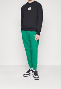 Sudadera negra con un parche de logo blanco, combinada con pantalones de chándal verdes que presentan un pequeño logo de la marca en blanco. Zapatillas deportivas en blanco y negro.