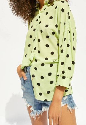 Femme portant une chemise à manches longues vert clair à boutons avec de gros pois noirs et un short en jean bleu déchiré.