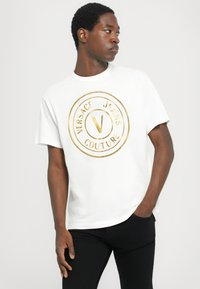 Balta kokvilnas T-kreklu ar lielu zelta apļa logo, kurā ir uzraksts "VERSACE JEANS COUTURE". Īsas piedurknes un apaļš kakla izgriezums.