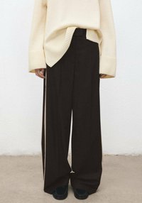 Pantalon large en tissu brun foncé avec des bandes latérales verticales, associé à un pull en maille beige clair avec des poignets côtelés.