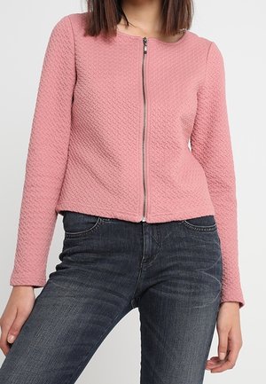 Vrouw draagt een lichtroze zip-up jack met een getextureerd oppervlak en lange mouwen, gecombineerd met donkergrijze strakke jeans, tegen een effen achtergrond.