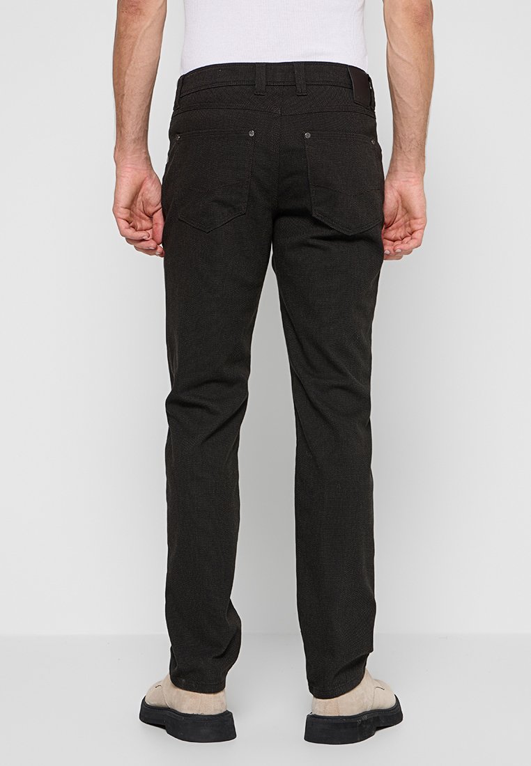 Mann in schwarzen Slim-Fit-Hosen und beigen Schuhen, der mit dem Rücken zur Kamera vor einem schlichten grauen Hintergrund steht.