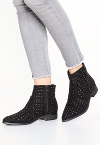 Botas de tobillo de ante negro con acentos plateados en forma de tachuelas, punta afilada y cremallera lateral, combinadas con jeans grises de dobladillo deshilachado.