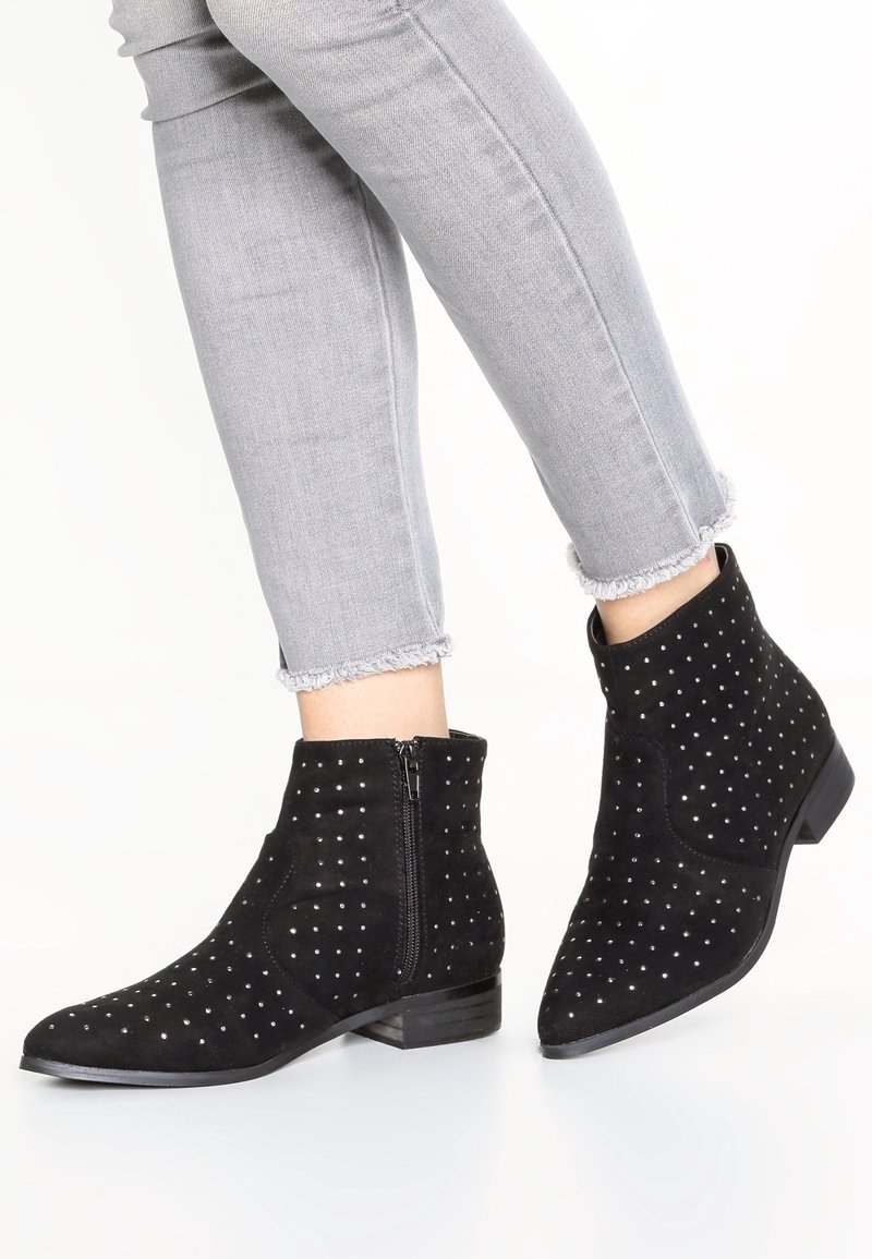 Botas de tobillo de ante negro con acentos plateados en forma de tachuelas, punta afilada y cremallera lateral, combinadas con jeans grises de dobladillo deshilachado.