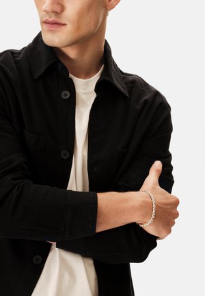 Silbernes Armband am Handgelenk mit einem Gliederdesign, getragen über einem schwarzen Hemd mit Knöpfen und einem weißen T-Shirt. Schlichter und moderner Look.