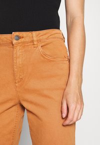 Vaqueros de mezclilla naranja con cierre frontal de botón, cinco bolsillos y costuras visibles. Tejido texturizado con un diseño de pierna recta.