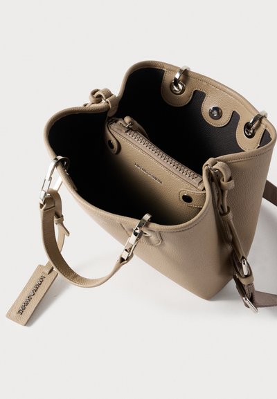 Μπεζ τσάντα κάδου με λείο δέρμα, ασημένια μεταλλικά στοιχεία, εσωτερική τσέπη με φερμουάρ και διπλούς ιμάντες. Ανοιχτό πάνω μέρος με διακοσμητικά γκρομέτ.