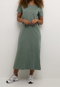 Sukienka t-shirt w oliwkowym kolorze z krótkimi rękawami, luźnym krojem i bocznymi rozcięciami, zestawiona z białymi i srebrnymi trampkami. Prosty, casualowy design.