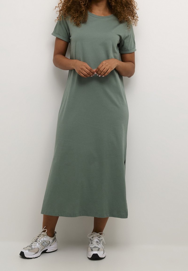 Sukienka t-shirt w oliwkowym kolorze z krótkimi rękawami, luźnym krojem i bocznymi rozcięciami, zestawiona z białymi i srebrnymi trampkami. Prosty, casualowy design.