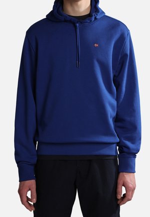 Hombre vistiendo una sudadera con capucha azul real con cordones y un pequeño emblema de la bandera noruega en el pecho, combinado con pantalones negros.